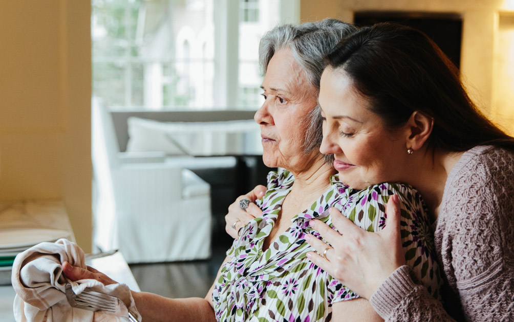 An older woman looks into the distance as her adult daughter hugs her around the shoulders.
