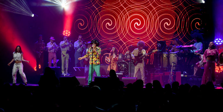 Several performers sing and play instruments onstage, with swirling lights projected behind them.