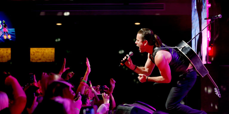 Charles Esten sings into a microphone as the audience puts their hands in the air.