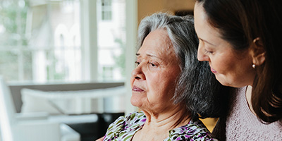 An older Hispanic/Latina woman stares off into space while a younger woman looks at her kindly.