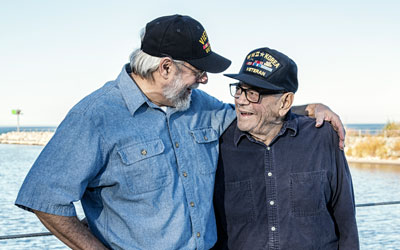 An older man who is wearing a Vietnam War veteran hat smiles and stands with his arm around an older man who is wearing a WW2 and Korean War veteran hat.